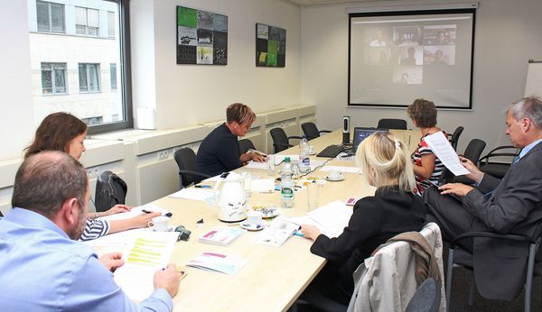 Foto zeigt Mitglieder der Jury an einem Konferenztisch. Über eine Videokonferenz sind weitere Jury-Mitglieder live dabei