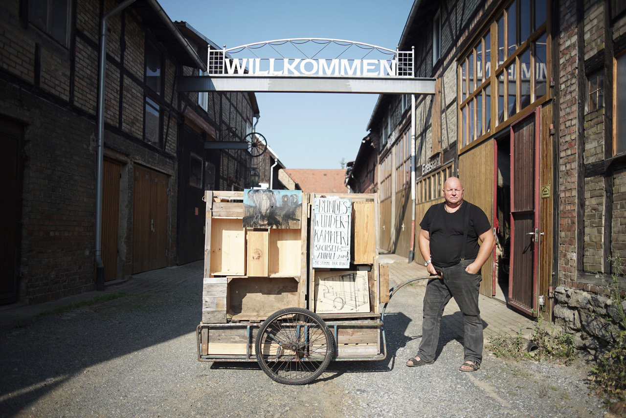 Künstler und Wundersammler Marc Haselbach steht mit seinem leeren Wunderwagen in einer Gasse zwischen Backsteinhäusern.
